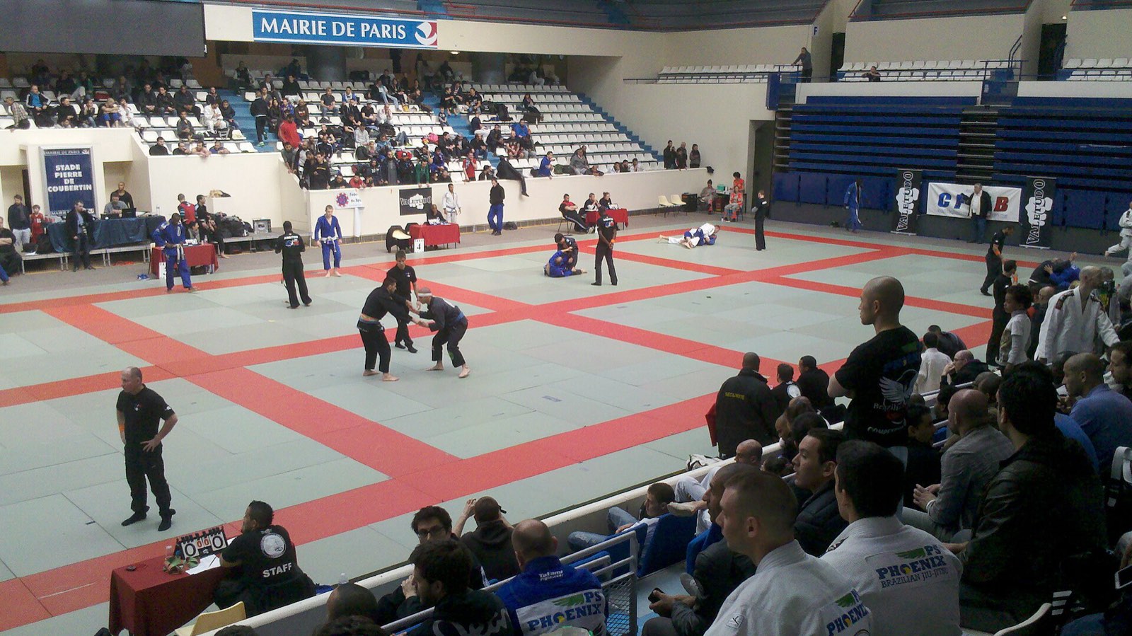 Arena de competição com vários tatames de jiu-jitsu rodando chaves em paralelo